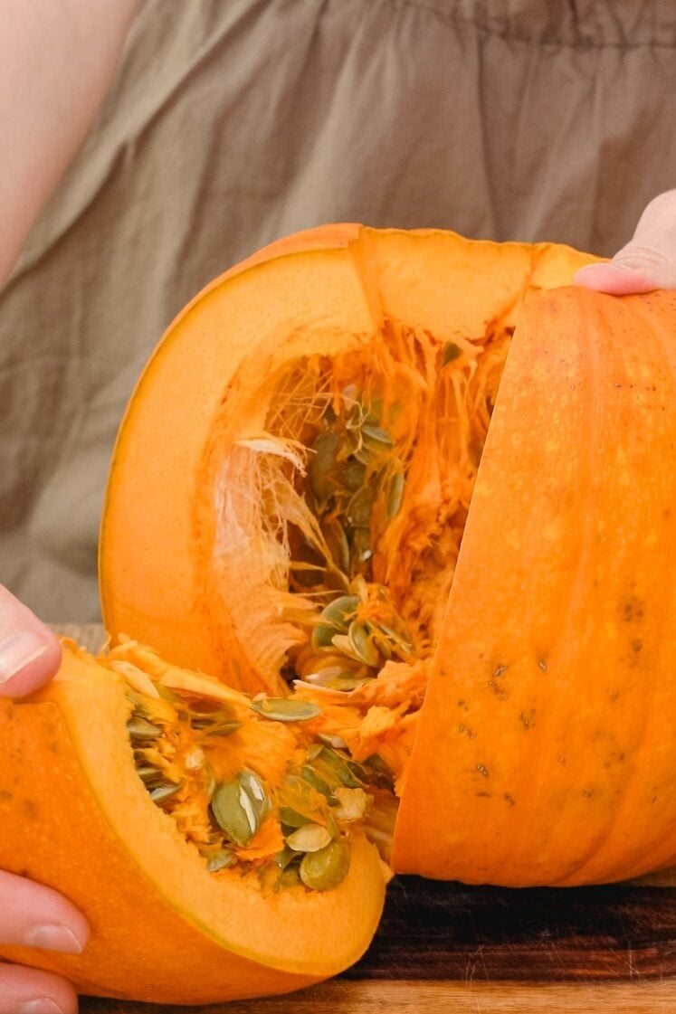 A person is holding a halved pumpkin, revealing its seeds and stringy insides. The pumpkin is bright orange, and the person is wearing a brown shirt.