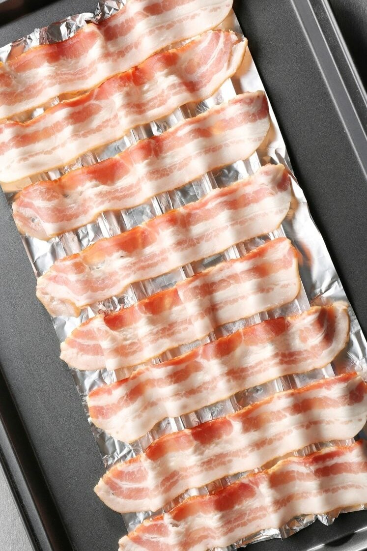 Several strips of raw bacon are laid out in a single layer on a baking sheet lined with aluminum foil, ready for cooking. The bacon shows a mix of pink meat and white fat, arranged neatly in parallel rows.