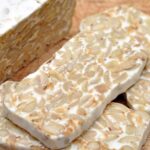 Slices of tempeh, a fermented soy product, are placed on a wooden surface. The tempeh has a firm texture with visible soybeans and a light, nutty appearance.