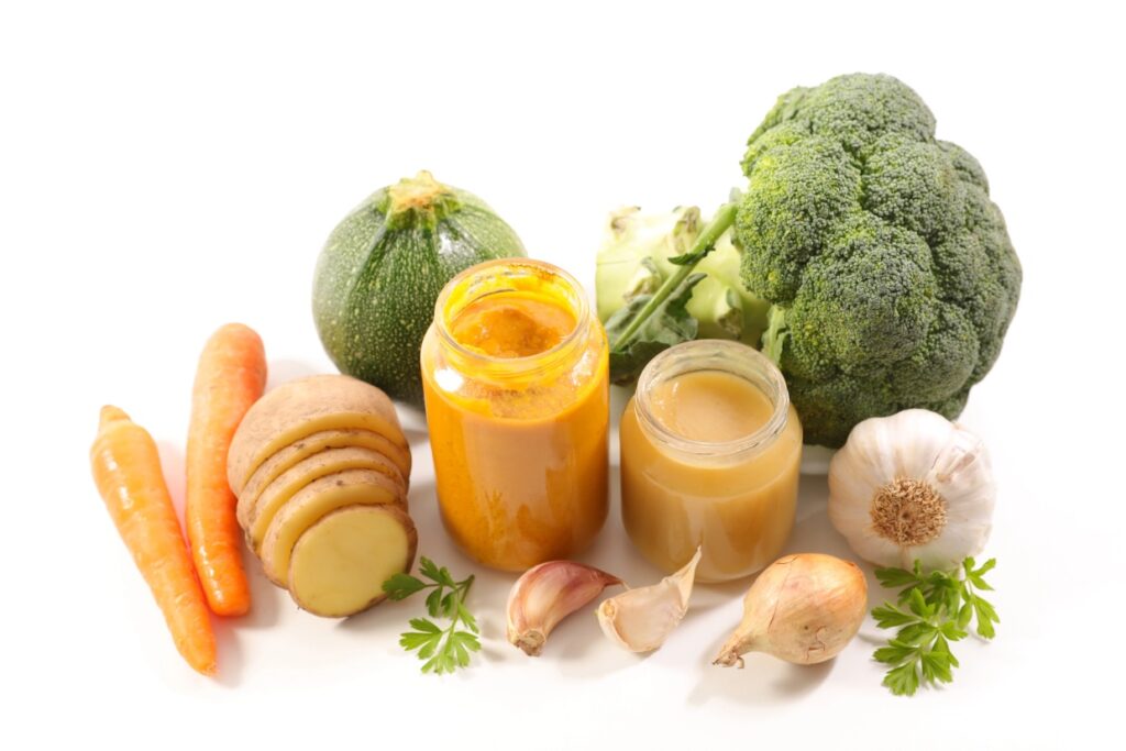 A display of fresh vegetables and spices, including a zucchini, broccoli, carrots, sliced ginger, garlic cloves, onions, and two open jars of sauce on a white background. Small parsley sprigs add a touch of green.