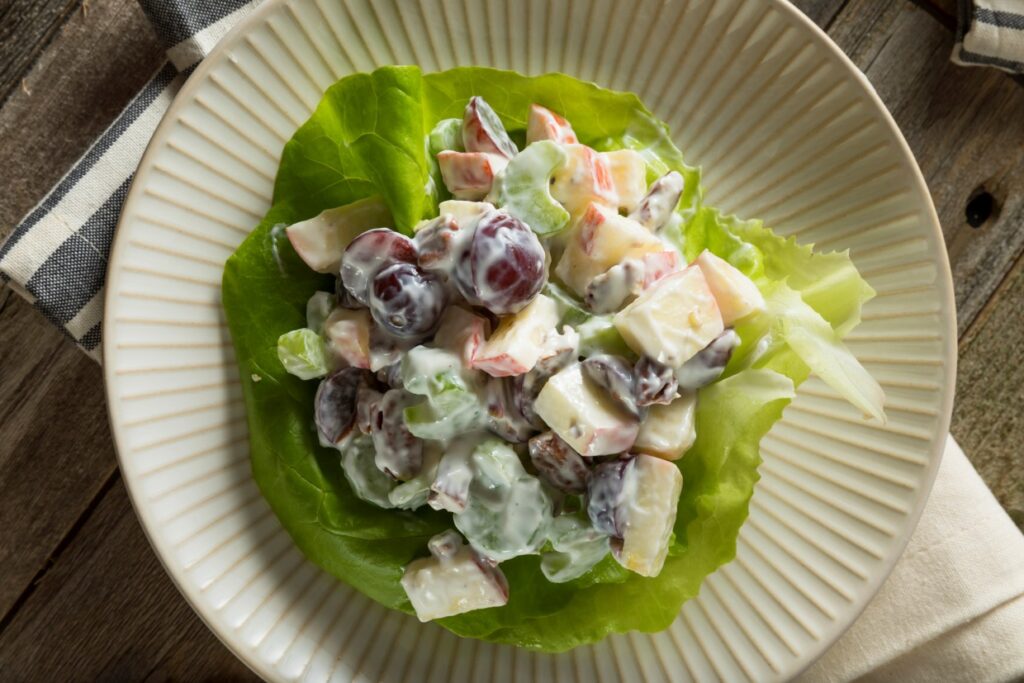 A close-up of a Waldorf salad on a light-colored plate. The salad includes diced apples, grapes, walnuts, and celery mixed in a creamy dressing, served on a bed of lettuce. The plate is on a wooden surface with a striped cloth nearby.