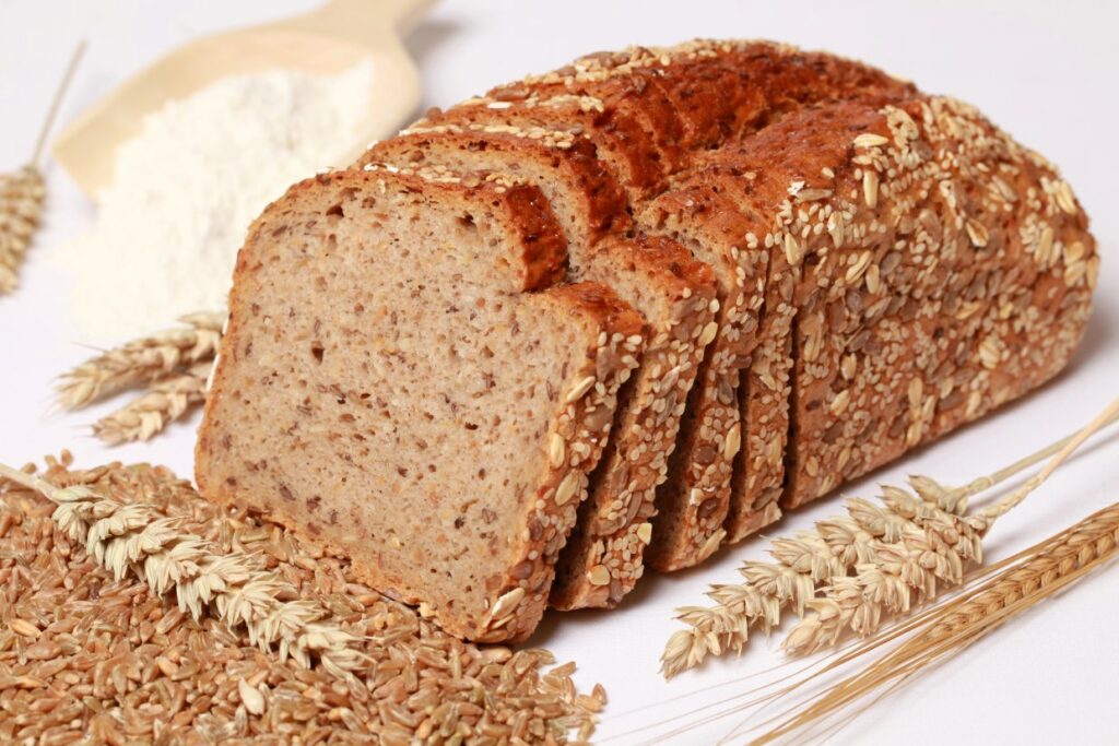 A loaf of whole grain bread partially sliced, topped with seeds. Surrounded by scattered wheat stalks and grains, with a wooden scoop of flour in the background on a white surface.