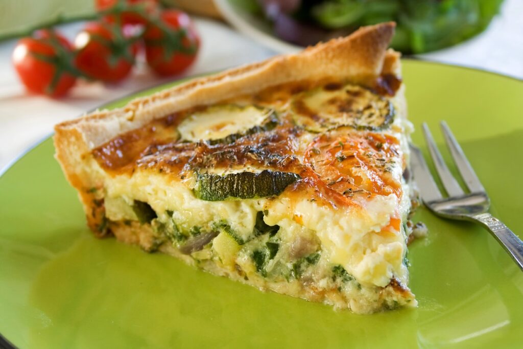 A slice of vegetable quiche on a green plate with a fork beside it. The quiche is filled with zucchini and other vegetables, with a golden-brown crust. In the background, there's a bunch of cherry tomatoes and leafy greens.