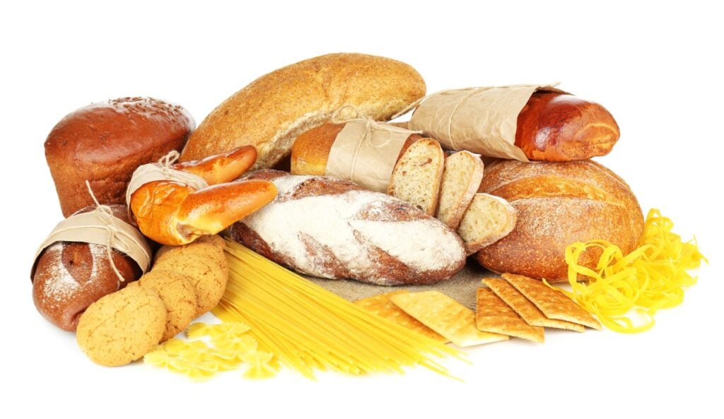 A variety of breads and pasta are displayed, including sliced and whole loaves, baguettes, cookies, and crackers. The pasta includes spaghetti and farfalle. Some items are wrapped in brown paper. The background is white.