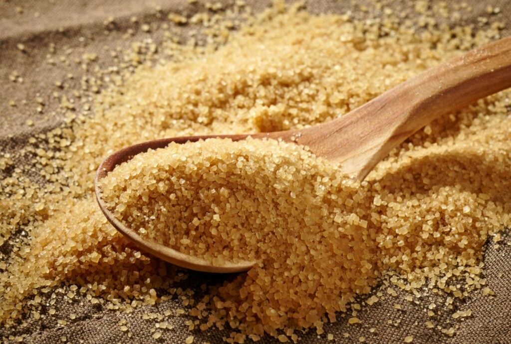 A wooden spoon scoops up coarse brown sugar from a mound on a textured fabric surface. The sugar grains glisten in the light, highlighting their rough texture.