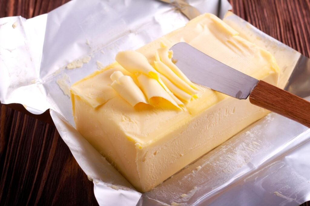 A block of butter partially unwrapped from foil sits on a wooden surface. A knife with a wooden handle rests on top, with curls of butter on its blade.