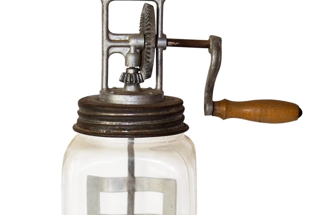 An antique glass butter churn with a wooden handle and metal gear mechanism. The clear jar has a metal lid with a crank on top, used for churning butter. The background is white.