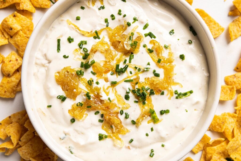 A bowl of creamy dip topped with caramelized onions and chopped chives, surrounded by corn chips.