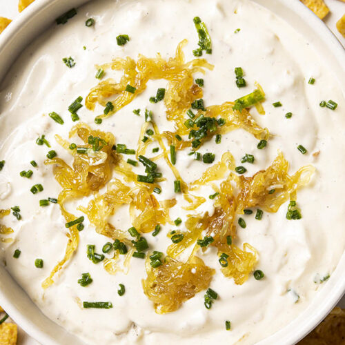 A bowl of caramelized onion dip garnished with caramelized onions and chopped chives, surrounded by crispy corn chips.