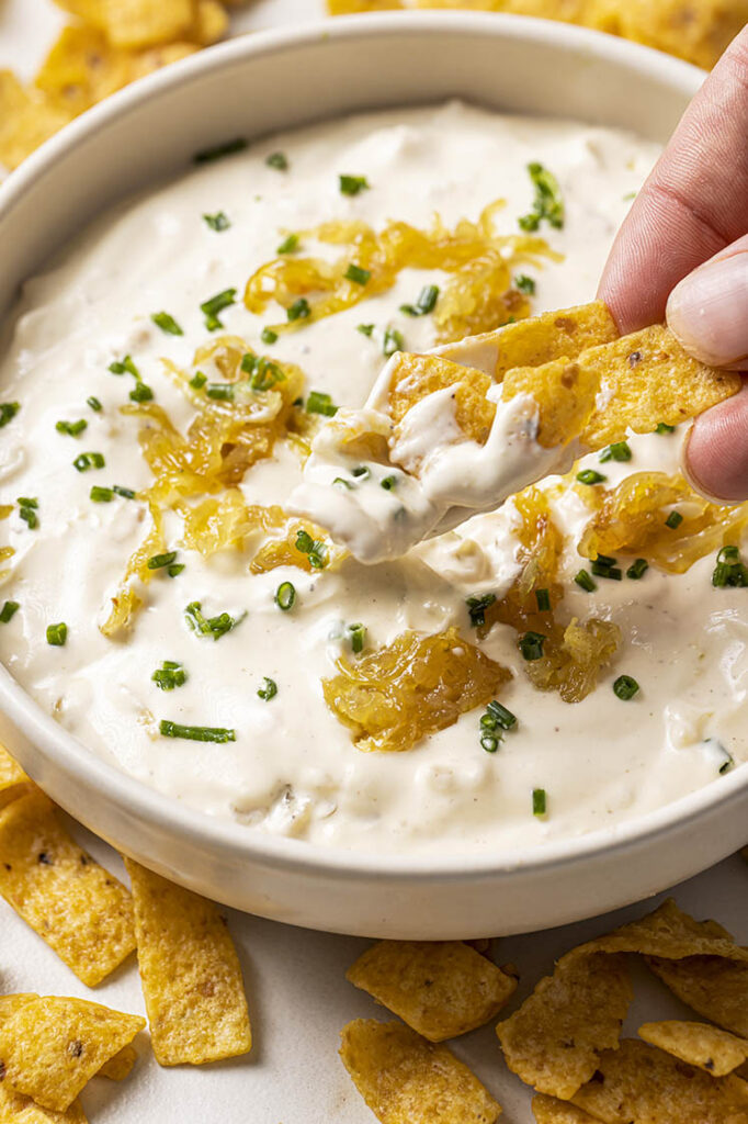 A hand dips a chip into the caramelized onion dip topped with caramelized onions and chopped chives. The bowl is surrounded by more chips.