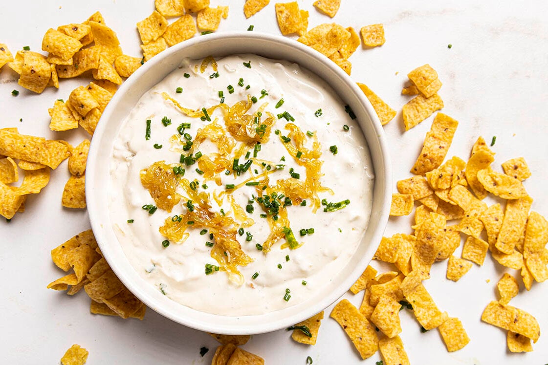 A bowl of caramelized onion dip garnished with caramelized onions and chopped chives, surrounded by crispy corn chips.
