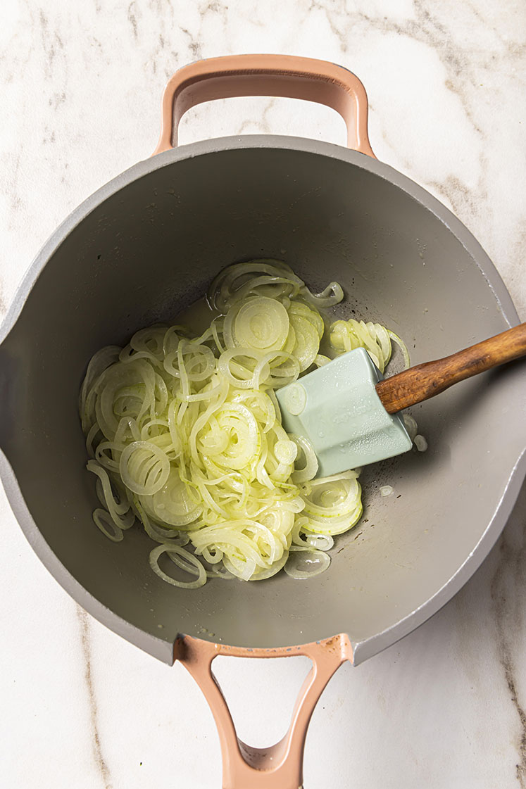 A pot with thinly sliced onions being sautéed, stirred with a light blue spatula with a wooden handle.