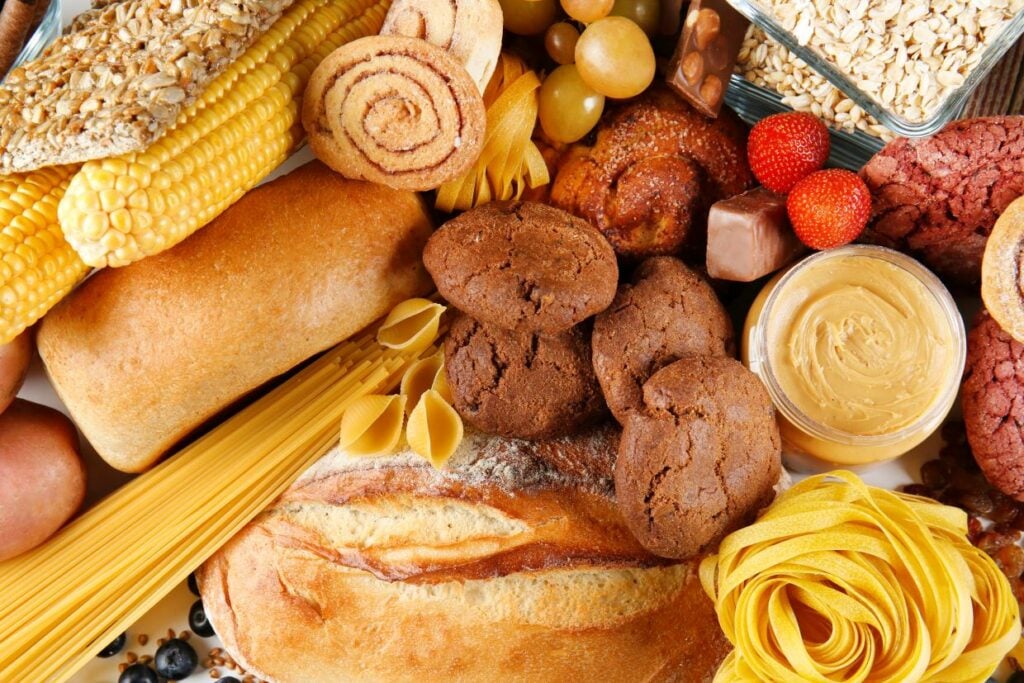 A variety of carbohydrate-rich foods, including bread, pasta, corn, cookies, pastries, oatmeal, strawberries, grapes, and peanut butter, are displayed. The image showcases a diverse selection of grains and baked goods.