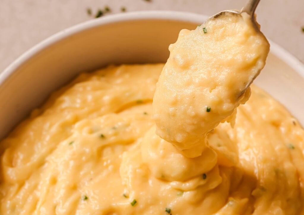 A close-up of creamy mashed potatoes in a white bowl, with a spoon lifting a portion. The mashed potatoes are smooth, with small green herbs sprinkled throughout, likely chives.