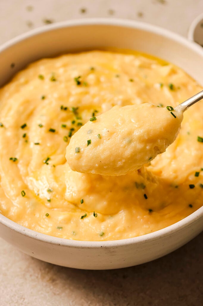 A spoon scoops cheddar and chive mashed potatoes from a bowl.