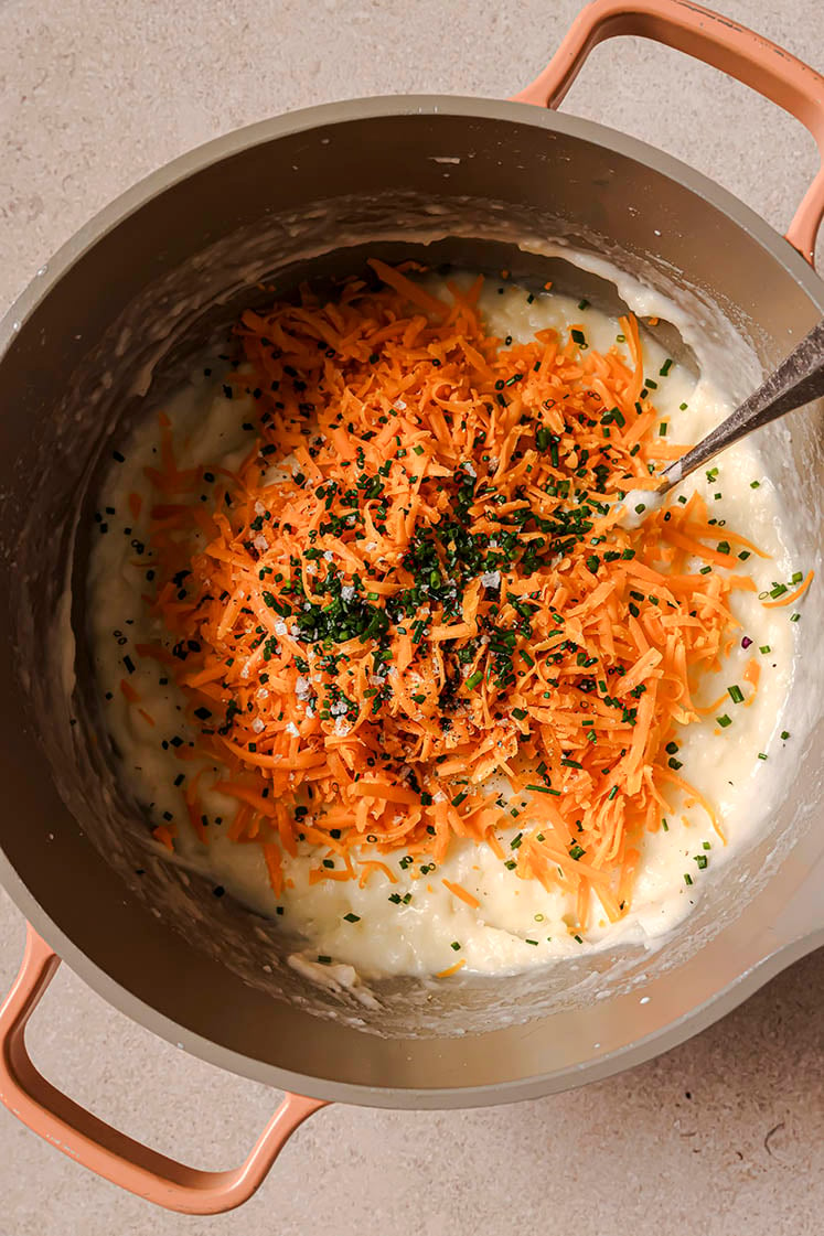 A pot of creamy mashed potatoes with shredded cheddar cheese, chopped chives, salt, and black pepper, with a spoon.