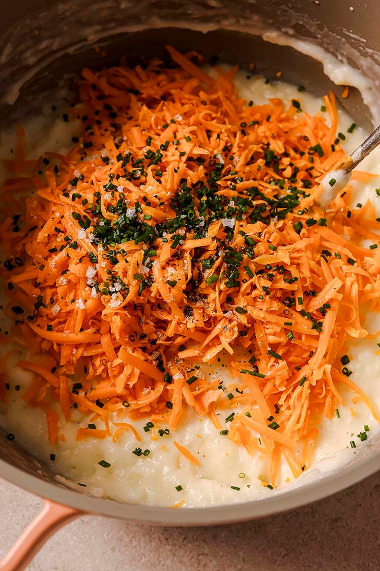 A pot of creamy mashed potatoes with shredded cheddar cheese, chopped chives, salt, and black pepper, with a spoon.