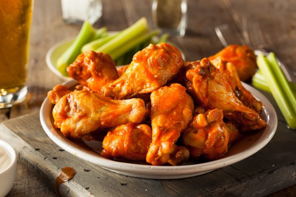 A plate of spicy buffalo chicken wings is piled high. The wings are glazed with a bright orange sauce. In the background, there are celery sticks and a glass filled with a yellow beverage on a rustic wooden table.