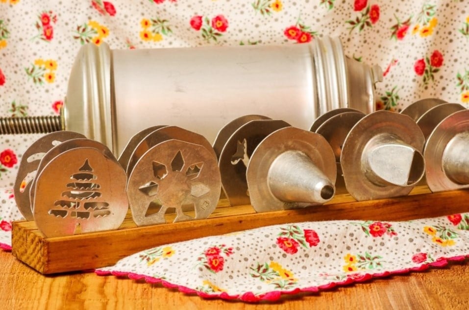 A vintage cookie press with multiple metal discs and a wooden stand is displayed. The discs have various cutout designs, including a Christmas tree and a snowflake. The background features a floral-patterned fabric.