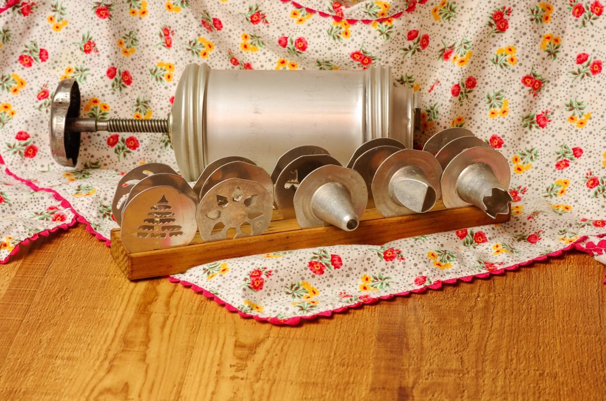 A vintage cookie press with multiple metal pattern disks on a wooden holder. The disks feature designs such as a Christmas tree and a flower. The setup rests on a floral, ruffled fabric atop a wooden surface.