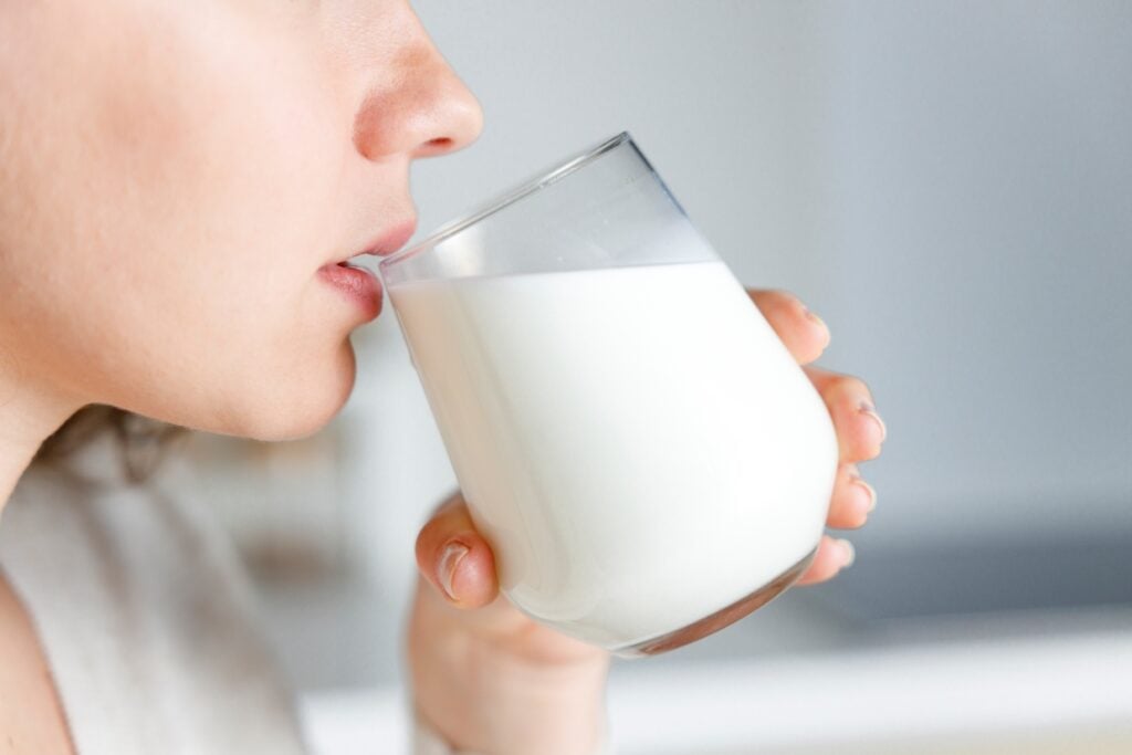 Close-up of a person drinking from a clear glass filled with milk. The focus is on the hand holding the glass and the mouth close to the rim. The background is softly blurred, creating a serene and simple atmosphere.