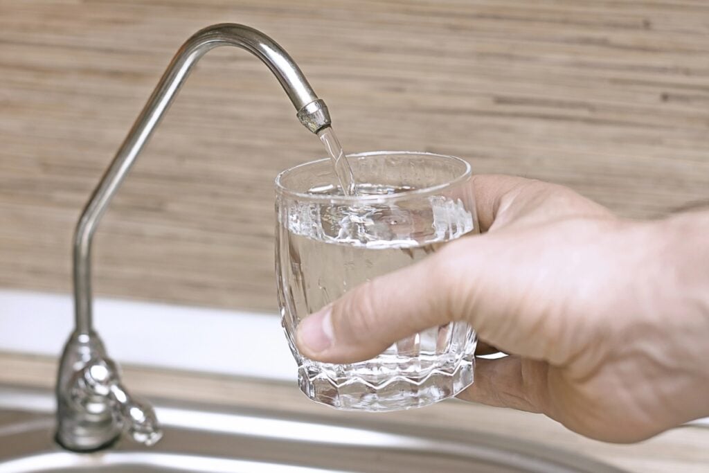 A hand holding a glass is being filled with water from a kitchen faucet. The background features a wooden-textured wall and a portion of a steel sink.