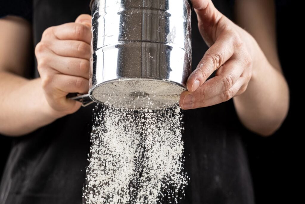 A person sifting flour through a metal sieve, with the flour falling in a fine stream. They are wearing a black apron, and the background is dark, highlighting the action of sifting.