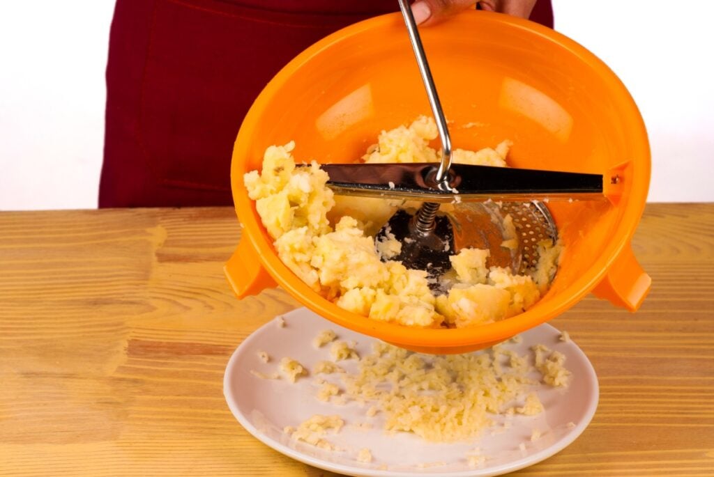 A person using a food mill to mash potatoes over a white plate. The orange bowl and stainless steel mill hold the potatoes, and the process is taking place on a wooden surface.