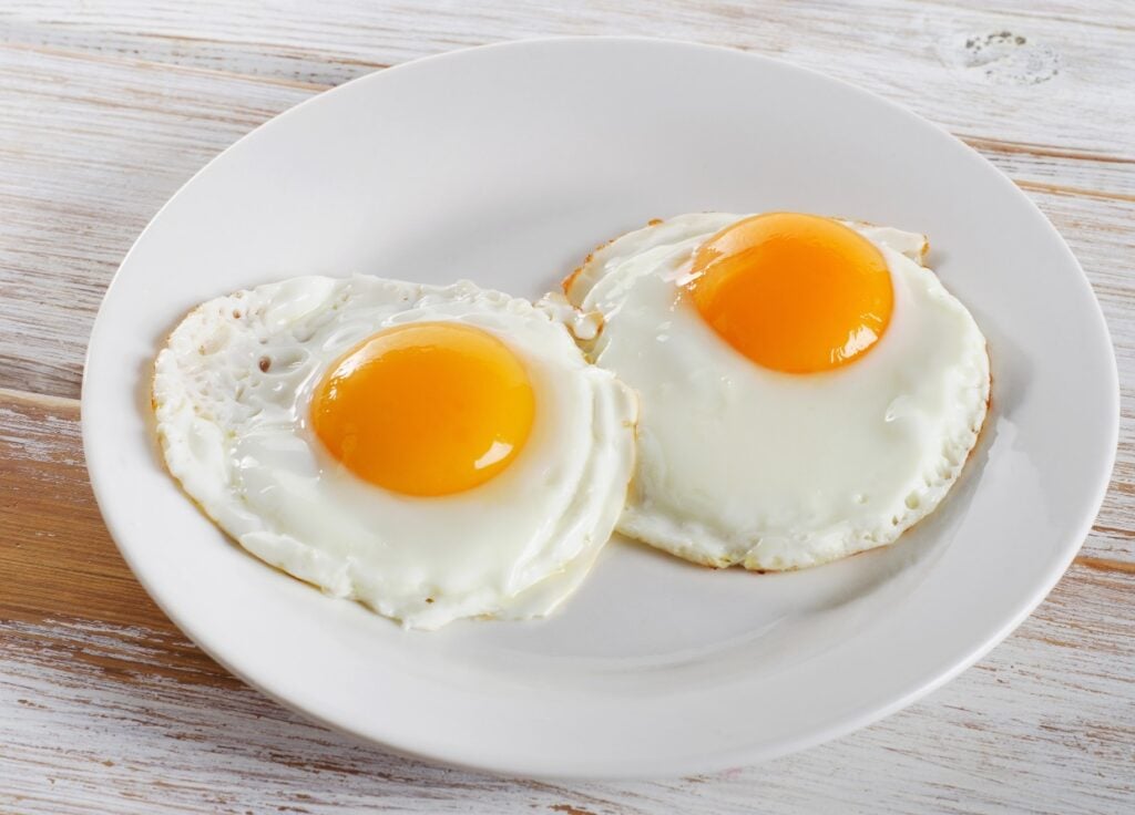 A white plate with two sunny-side-up eggs on a light wooden table. The eggs have bright yellow yolks and slightly crispy edges, arranged side by side.