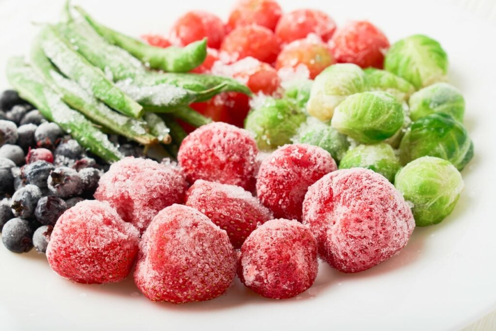 A plate featuring a variety of frozen vegetables and fruits. There's a cluster of strawberries, green beans, blueberries, brussels sprouts, and cherry tomatoes, all covered in frost.