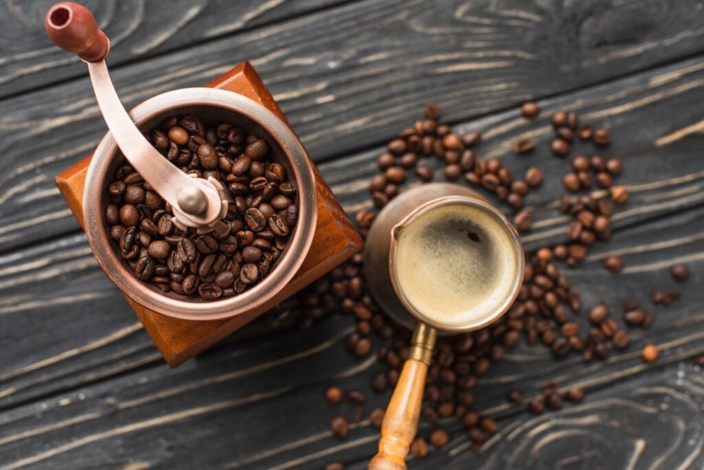 Top view of a manual coffee grinder filled with coffee beans next to a copper cezve with freshly brewed coffee. Coffee beans are scattered on a dark wooden surface, creating a cozy and rustic atmosphere.