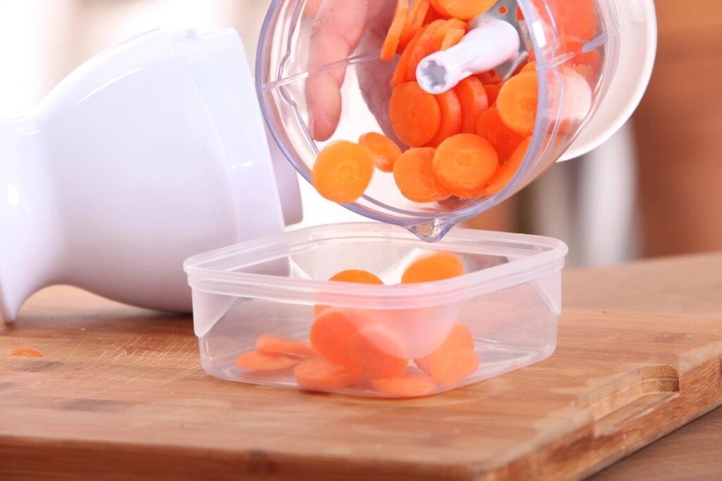 A person pours sliced carrots from a food processor into a clear plastic container on a wooden countertop.