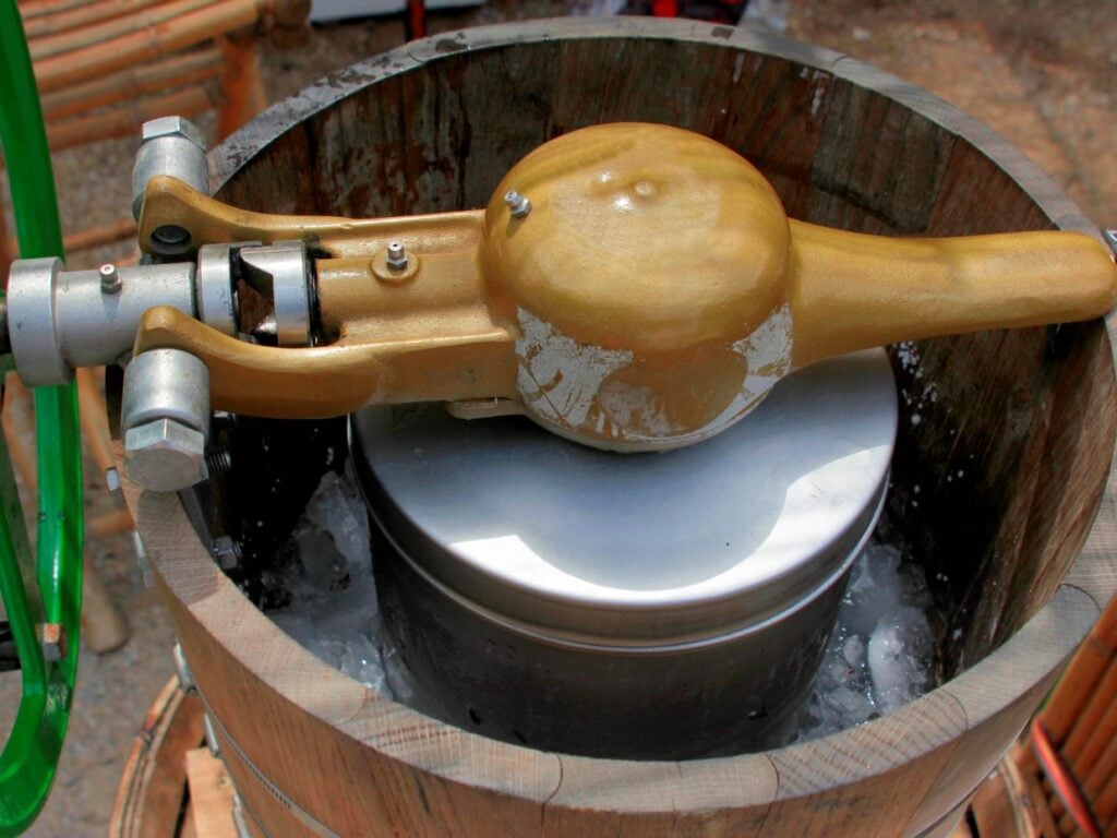 A vintage ice cream maker with a wooden barrel and metal hand crank mechanism is filled with ice. The device's top lid is golden and appears worn, indicating frequent use. The setting is outdoors, suggesting a homemade ice cream activity.