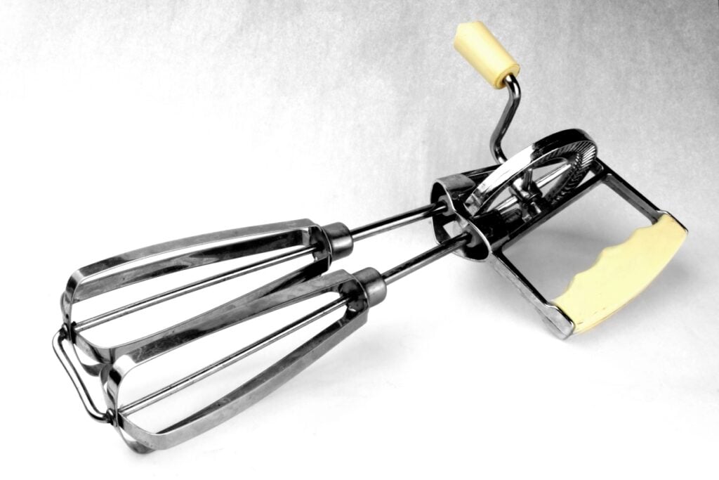 A vintage hand-crank mixer with metal beaters and a yellow handle, set against a white background.