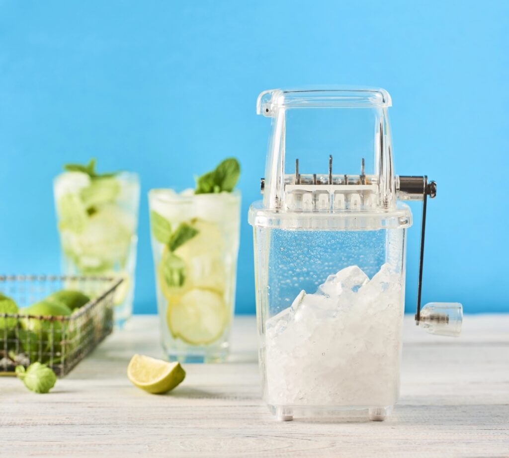 A clear manual ice crusher on a light wooden surface, filled with crushed ice. In the background are two mojito glasses garnished with mint leaves and a basket of limes. The scene is set against a bright blue background.