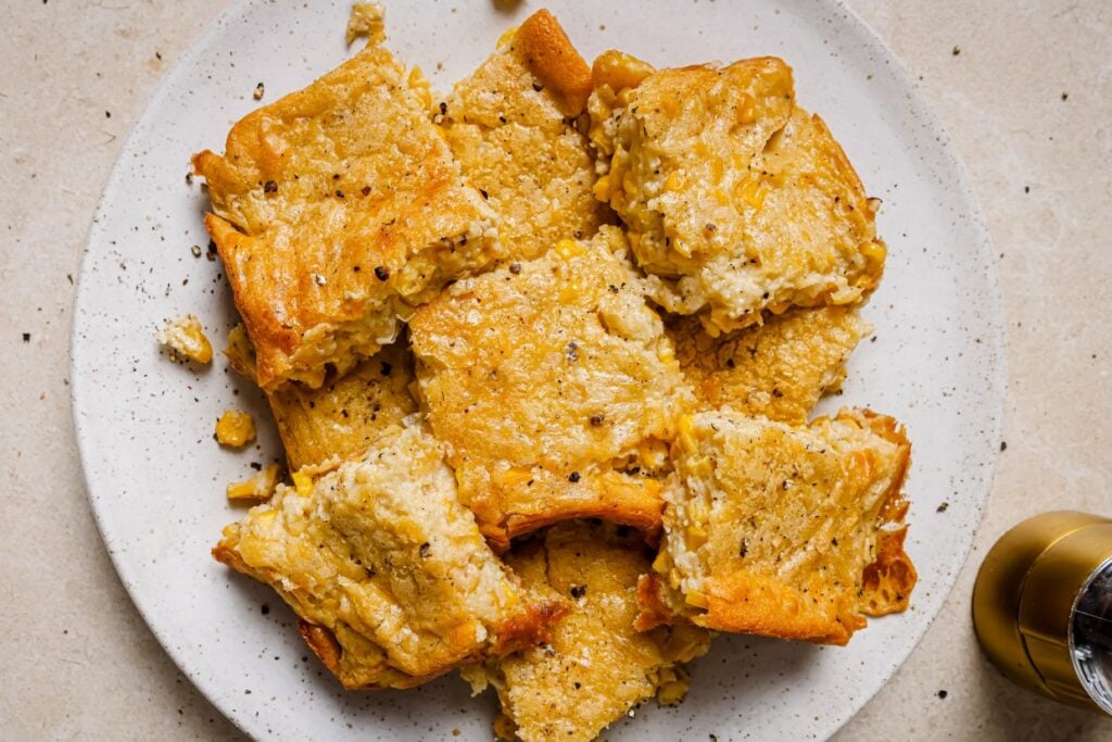 A white plate holds several slices of corn casserole topped with cracked black pepper. The slices are golden brown with a slightly crumbly texture, and a sprinkle of black pepper is visible on the creamy surface.