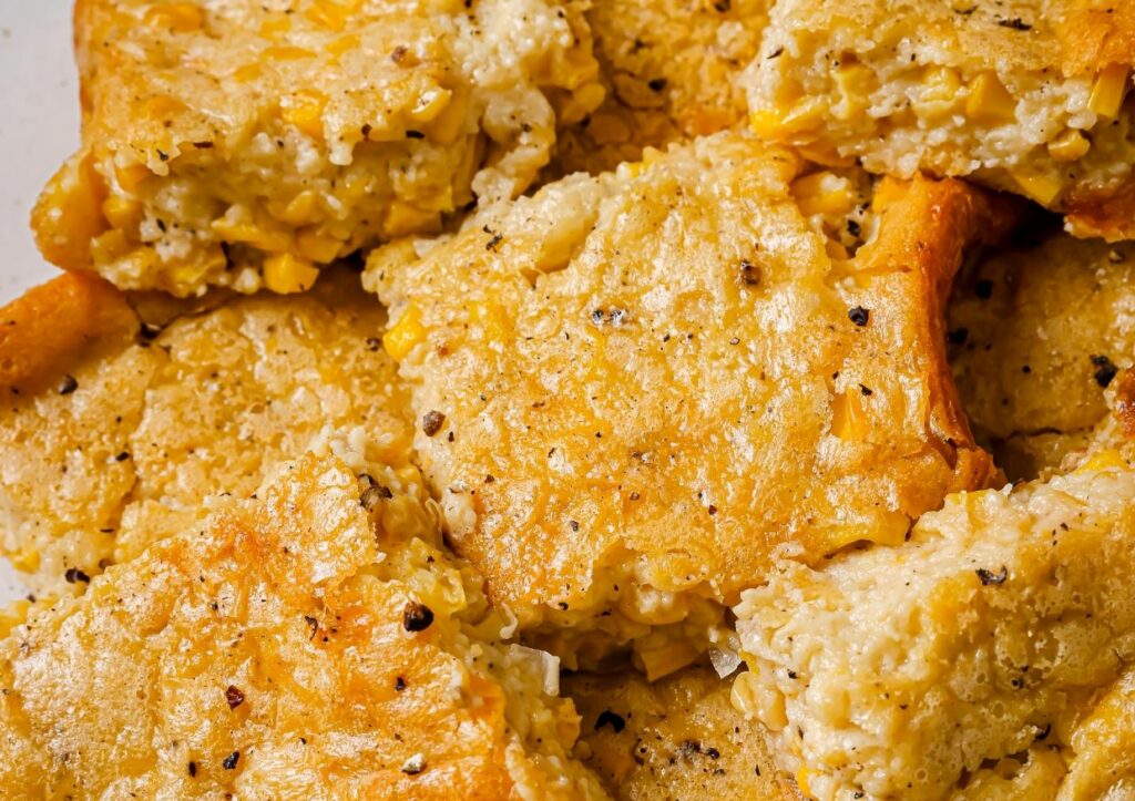 Close-up of several pieces of golden, crispy corn casserole topped with coarse black pepper. The texture is crumbly and moist, with visible corn kernels throughout.