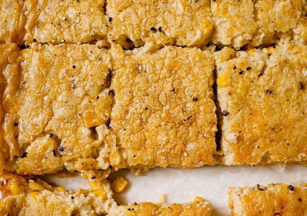 Close-up of baked cornbread cut into square pieces. The cornbread is golden brown with visible corn kernels and specks of black pepper, arranged closely together on a white surface.