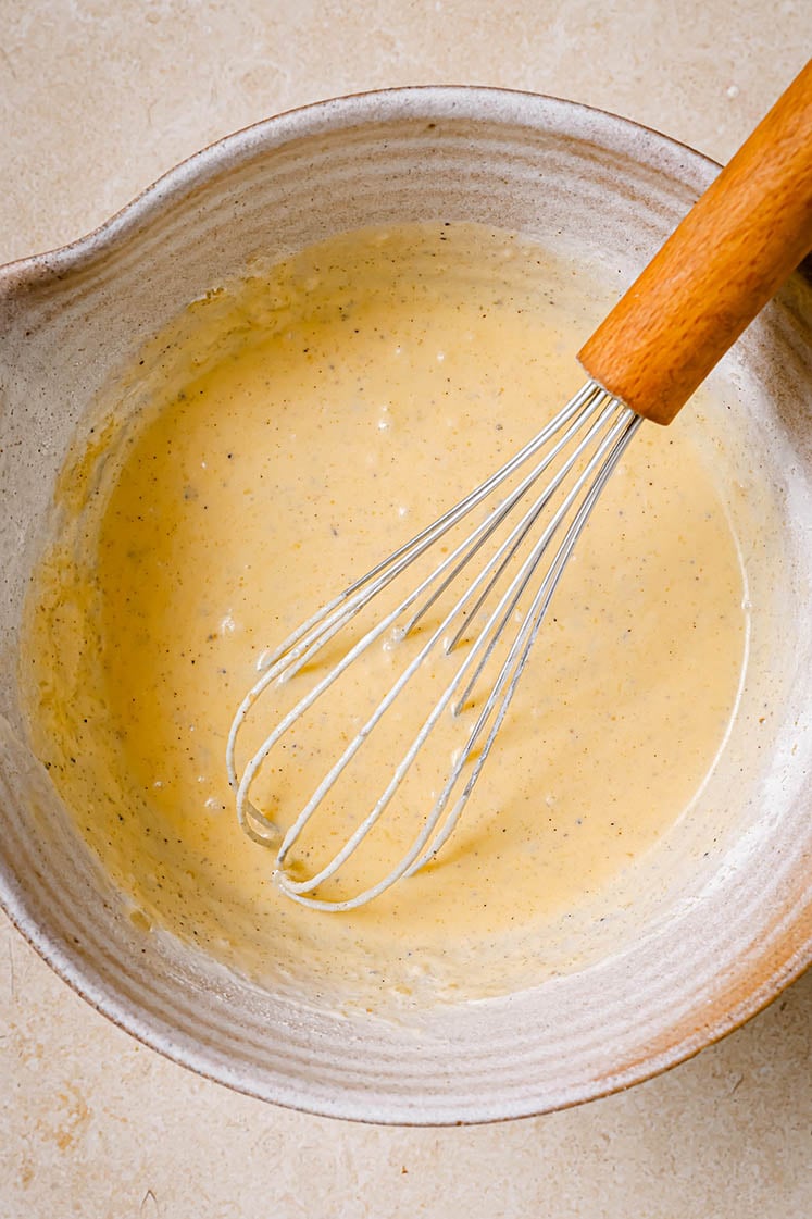 A ceramic bowl with eggs, sour cream, and melted butter being mixed by a whisk.