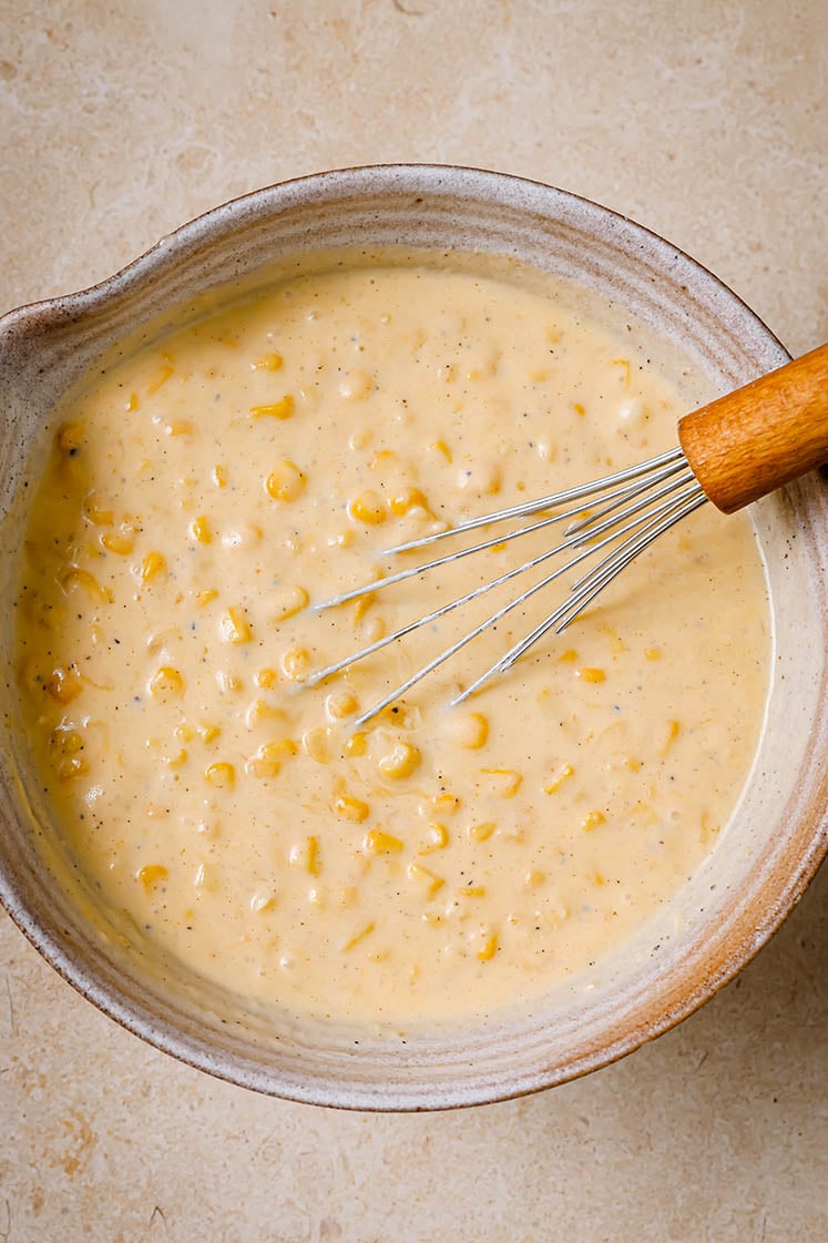 A ceramic bowl with Jiffy corn casserole batter being mixed by a whisk.