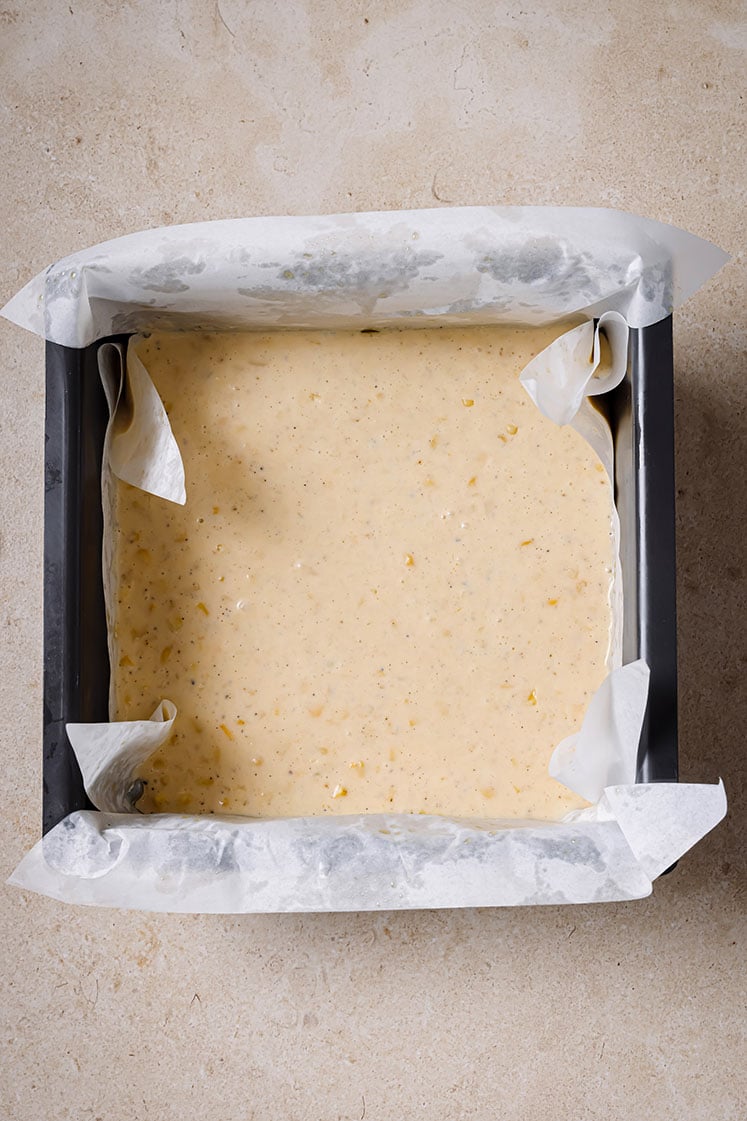 A square baking pan lined with parchment paper, filled with the Jiffy corn casserole batter, ready to be baked.