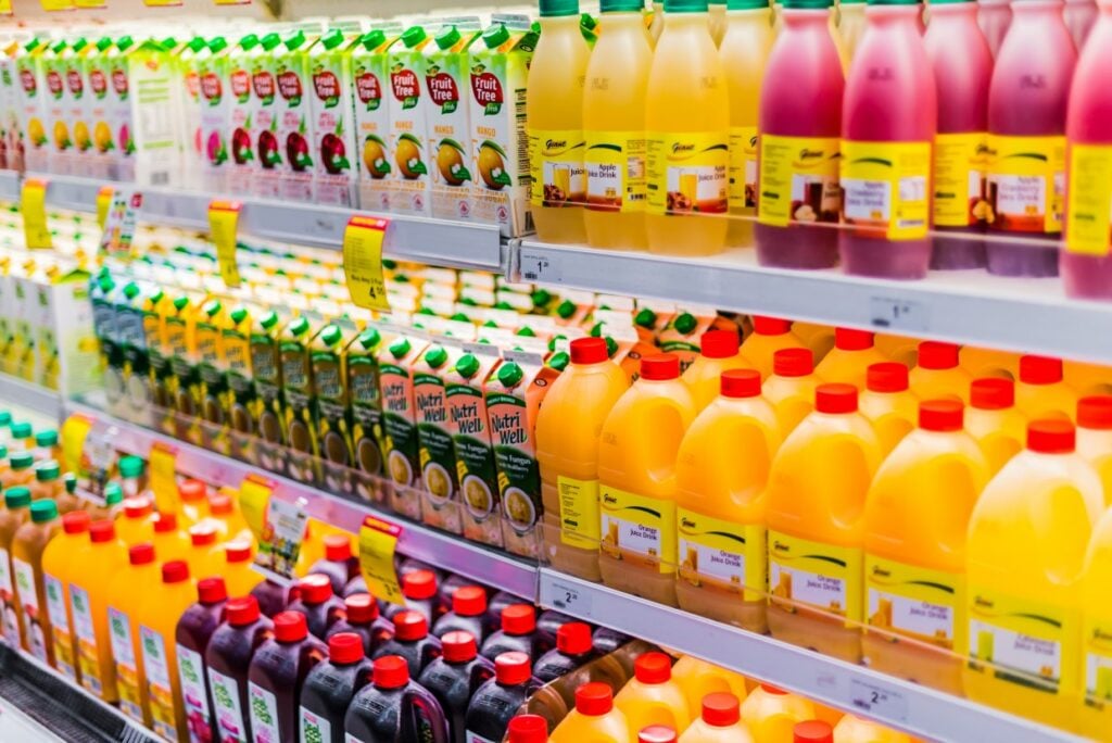 A supermarket shelf filled with a variety of colorful juice bottles and cartons. The selection includes orange juice, apple juice, and other fruit flavors, displayed in bright packaging and neatly organized in rows.