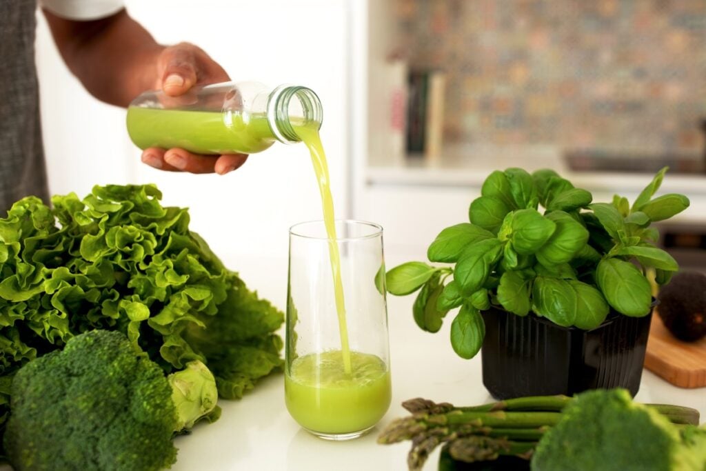 A person pours green juice from a bottle into a glass on a counter. Surrounding the glass are fresh vegetables, including lettuce, basil, broccoli, and asparagus, all arranged on a white surface.