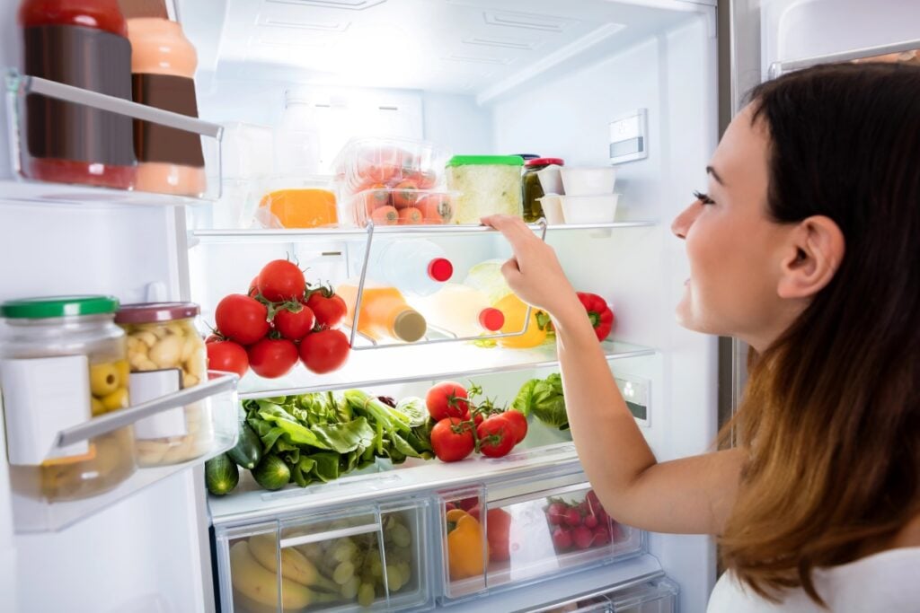 A woman reaches into a well-stocked refrigerator filled with fresh vegetables, fruits, and various containers. The shelves are neatly organized with tomatoes, greens, jars, and bottles in a brightly lit kitchen setting.