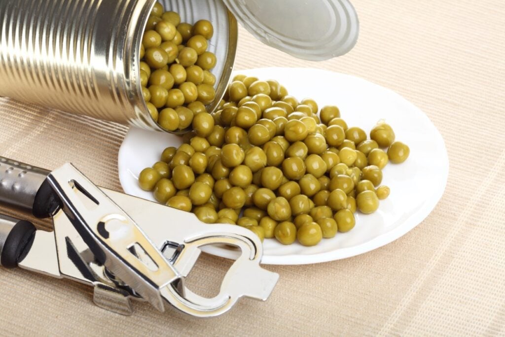 An opened can of green peas is tilted over a white plate, spilling peas onto it. A metal can opener is placed next to the can. The background is a neutral-toned textured surface.