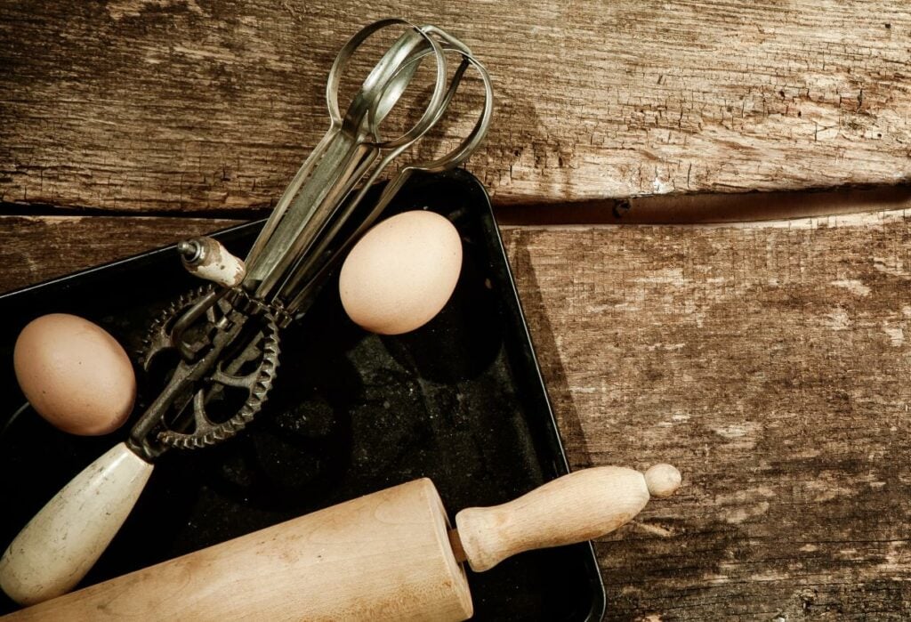 A vintage egg beater and two eggs rest in a baking tray on a rustic wooden surface. A wooden rolling pin is partially visible in the foreground, adding to the kitchen theme.