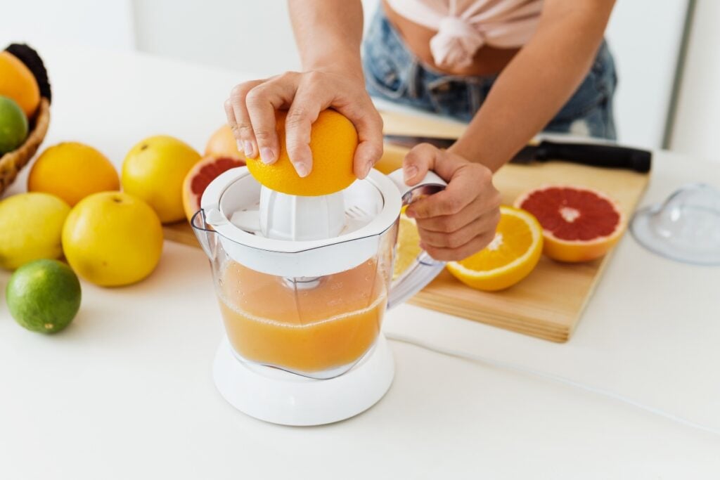 A person using an electric juicer to squeeze juice from an orange. Surrounding fruits, including grapefruits, limes, and oranges, are on the table. The juice collects in a clear container. A cutting board and knife are nearby.