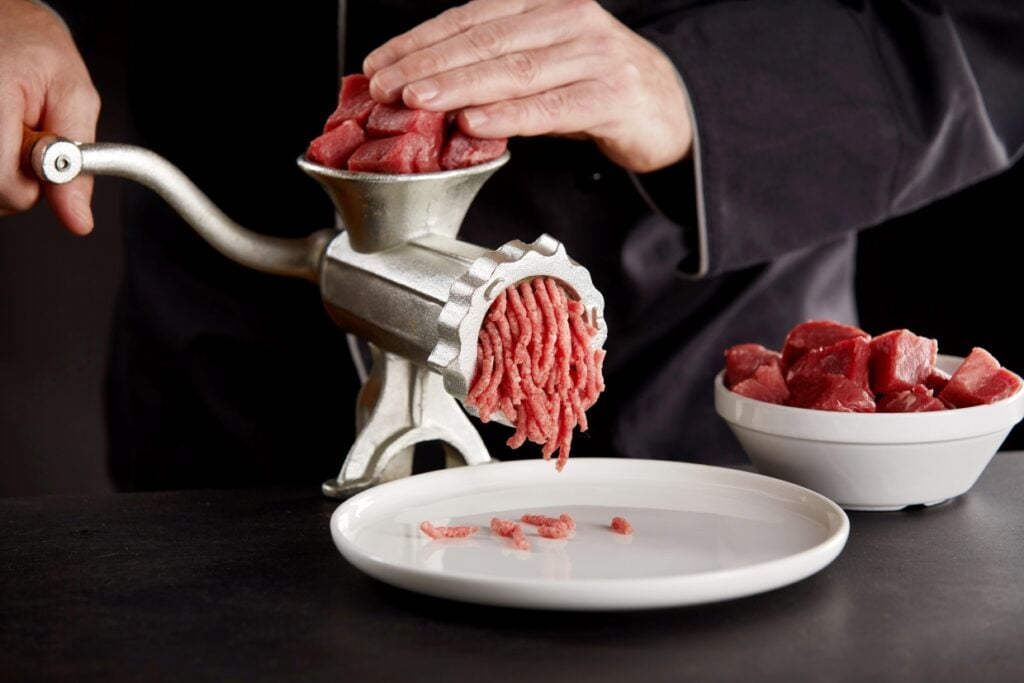 A person is using a metal meat grinder to grind pieces of raw beef into ground meat, which is falling onto a white plate. A bowl of cubed beef is beside the grinder on a dark countertop.