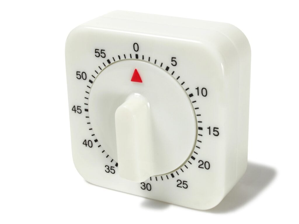 A white kitchen timer with a square shape and a round dial. The dial has black numbers and tick marks, ranging from 0 to 60. A red triangular pointer is set at 0. The timer has a white knob for setting the time.