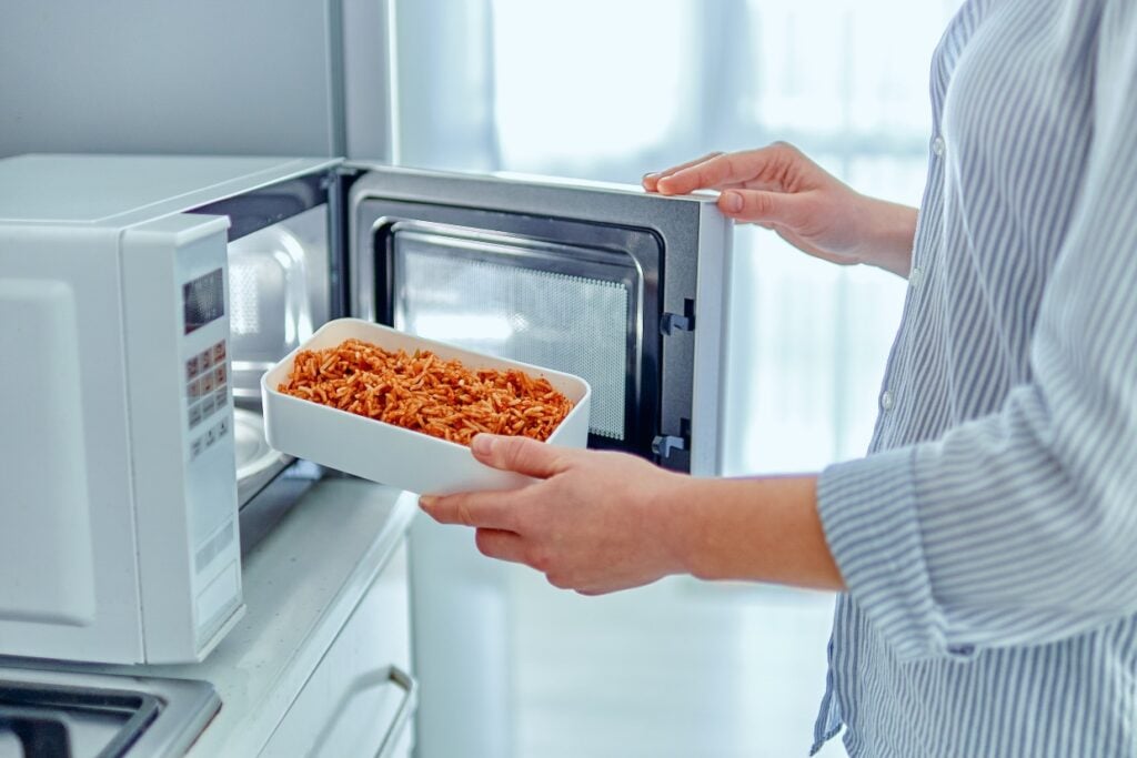 A person in a striped shirt is placing a white container with pasta into a microwave. The microwave door is open, and the kitchen setting appears bright and modern.